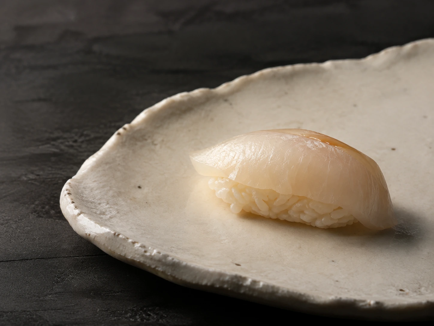 Um nigiri de peixe branco em cerâmica artesanal, iluminado de lado.